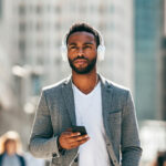 A man walking in an urban setting, wearing headphones and holding a smartphone, appearing focused as he listens to audio content. He is dressed in a casual yet stylish outfit, including a gray blazer and white T-shirt, with a blurred cityscape in the background.