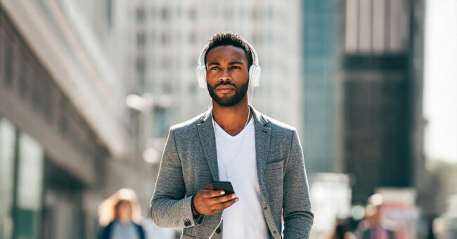 A man walking in an urban setting, wearing headphones and holding a smartphone, appearing focused as he listens to audio content. He is dressed in a casual yet stylish outfit, including a gray blazer and white T-shirt, with a blurred cityscape in the background.